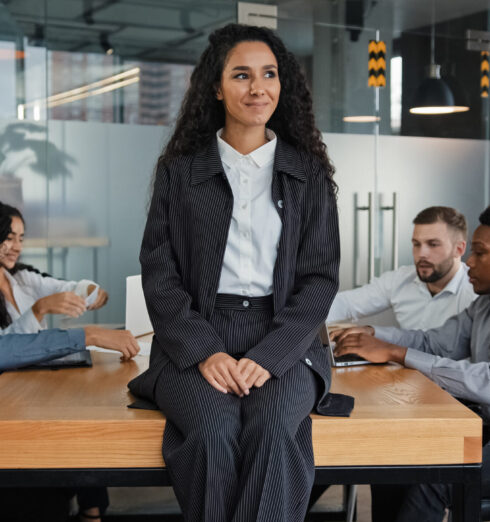 Pensive smiling businesswoman female leader boss woman sitting on table in office thinking. Multiracial employees multiethnic workers colleagues in background discuss work project working at meeting Pensive smiling businesswoman female leader boss woman sitting on table in office thinking. Multiracial employees multiethnic workers colleagues in background discuss work project working at meeting. High quality photo
